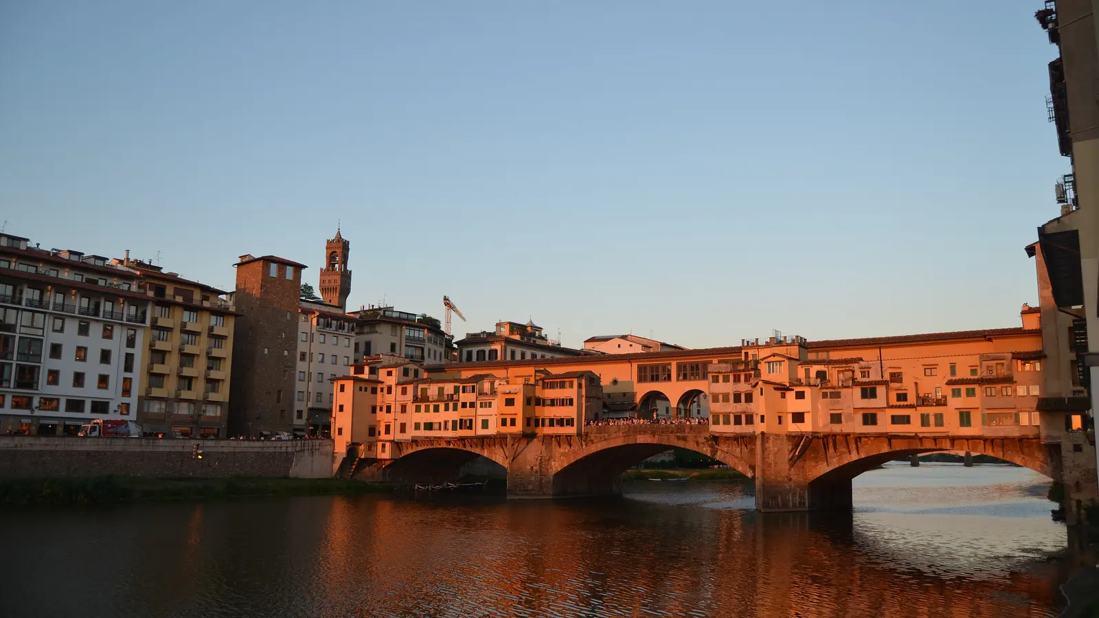 Der Ponte Vecchio bei Sonnenuntergang, seine Läden leuchten orange über dem Arno, dahinter der Turm des Palazzo Vecchio