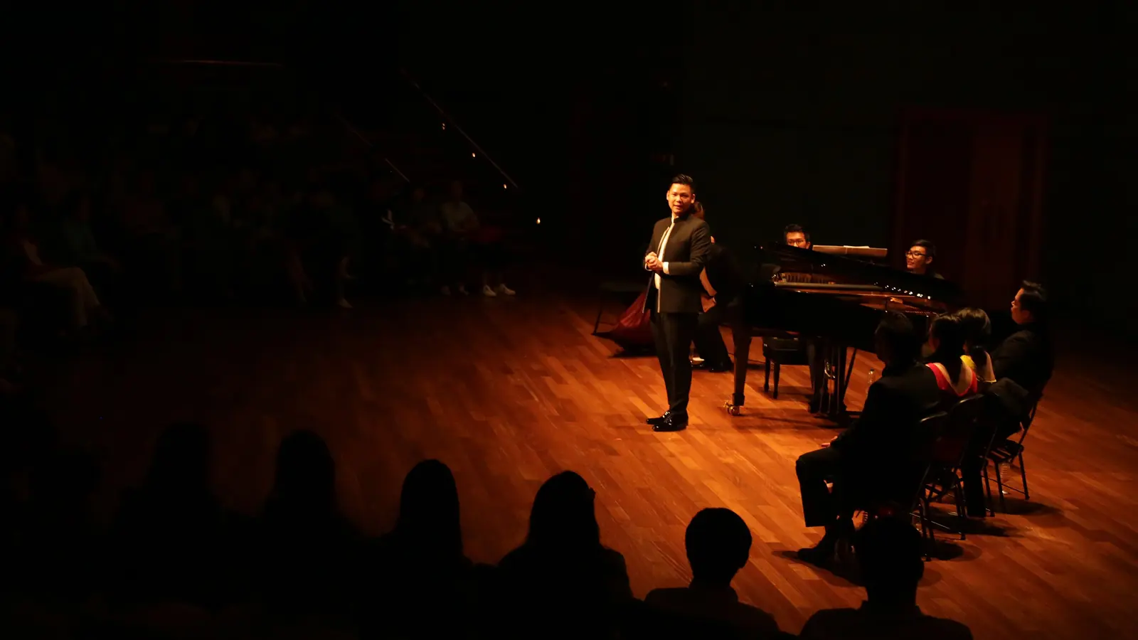 Ein Tenor steht im warmen Scheinwerferlicht neben einem Flügel, während ein dunkles Publikum lauscht; ein Tenorrezital in einem anderen Saal, nicht das Florenz-Konzert